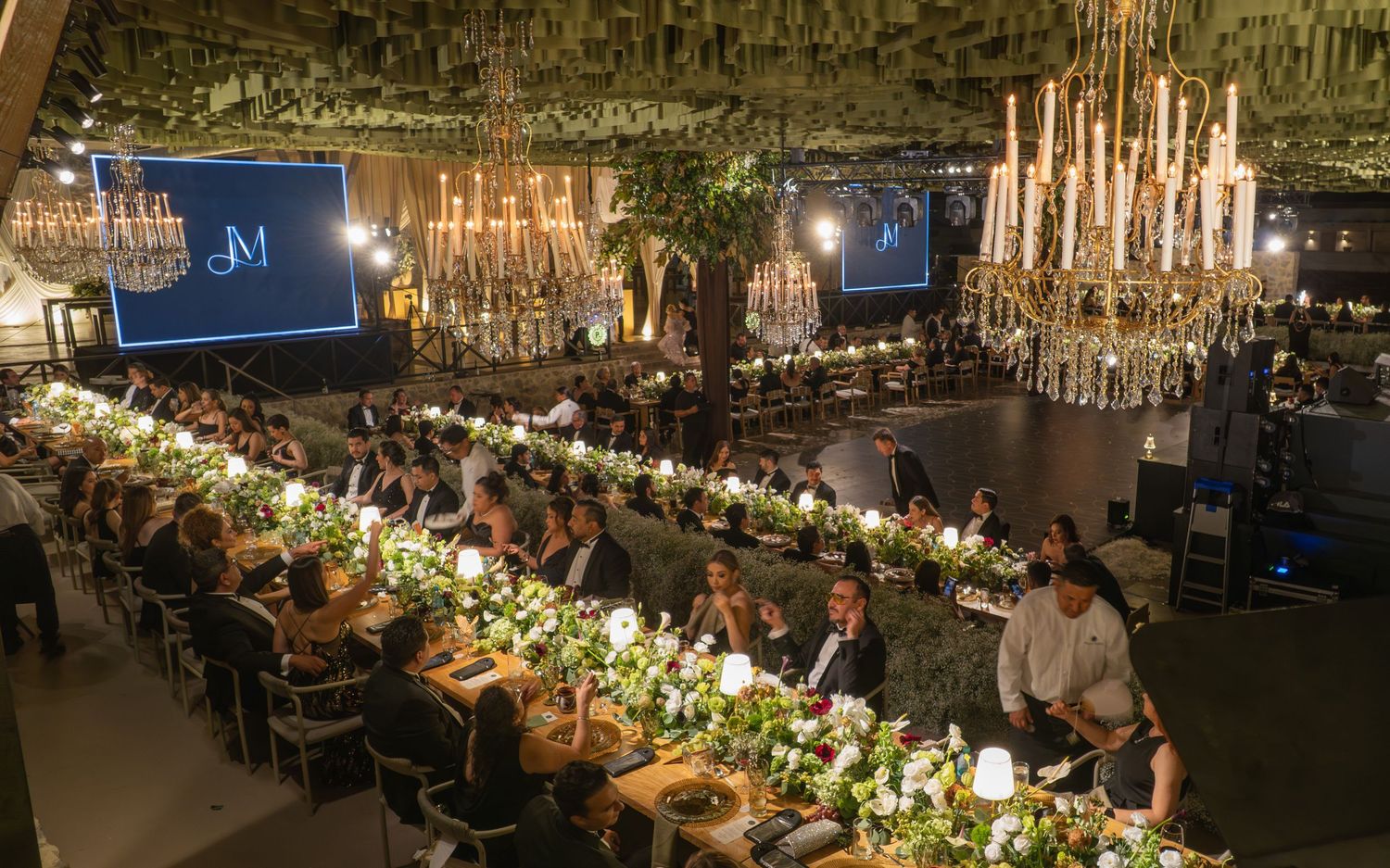 A long banquet table decorated with elegant chandeliers, floral arrangements, and place settings in a dimly lit venue.