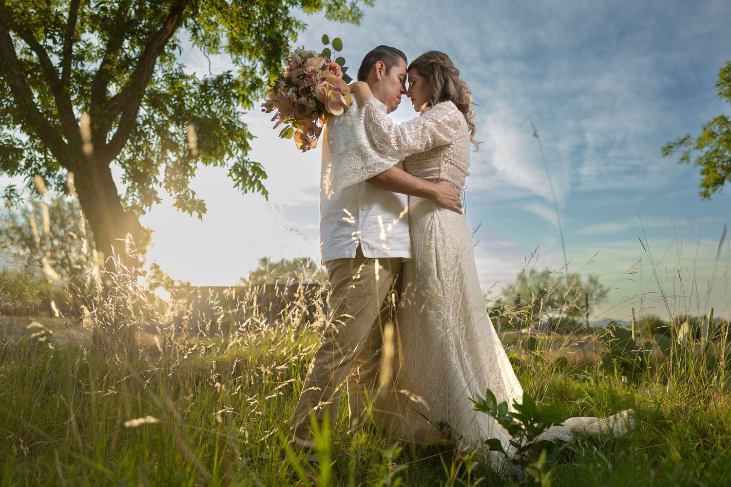 A couple embraces in tall grass during golden hour with a bouquet of flowers. Sesión en Parras Coahuila