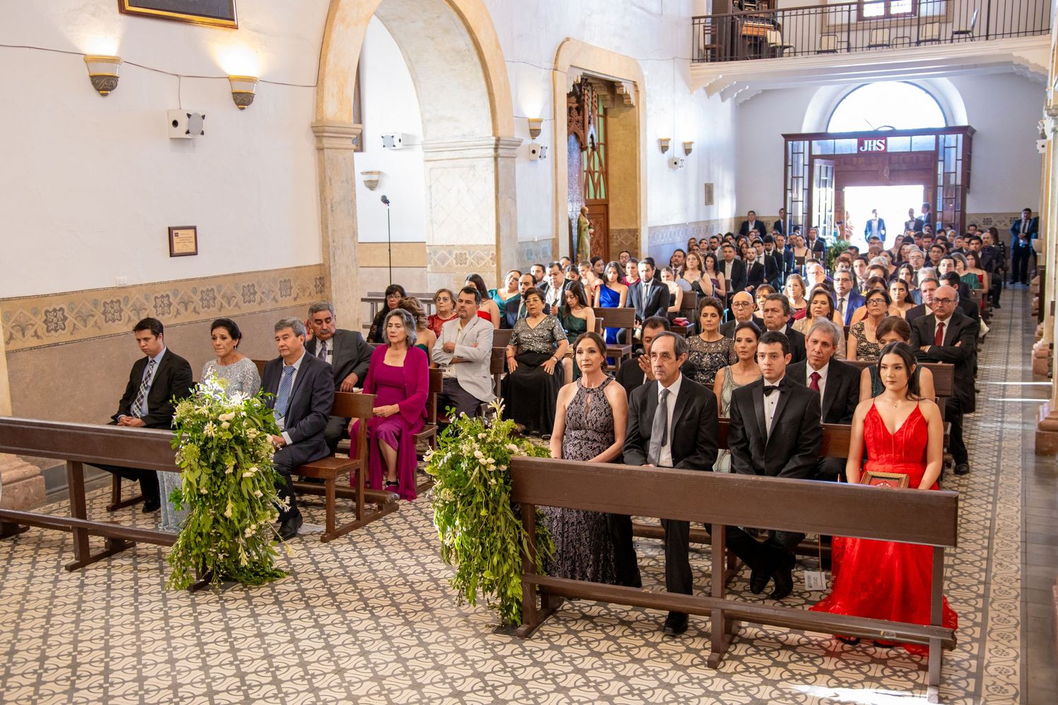 Wedding guests seated in church pews decorated with greenery during ceremony. Boda en Parras Coahuila, Fotógrafo de bodas
