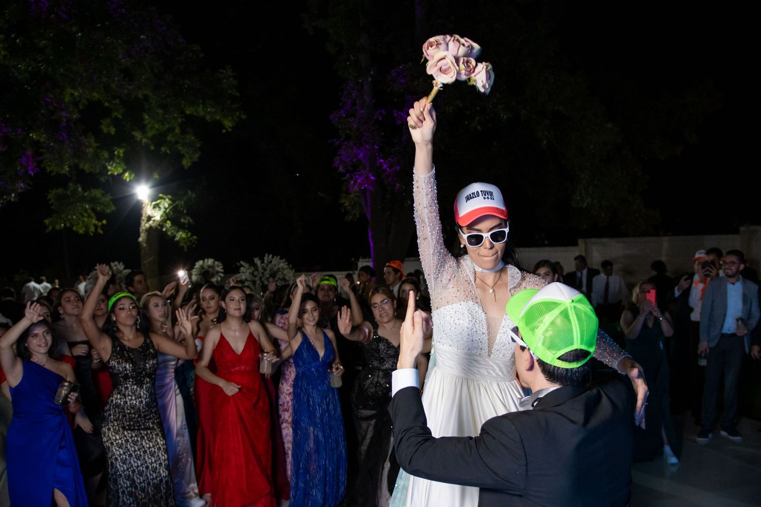 A person in white holds up a bouquet during a nighttime wedding celebration surrounded by cheering guests. Boda en Parras Coahuila, Foto