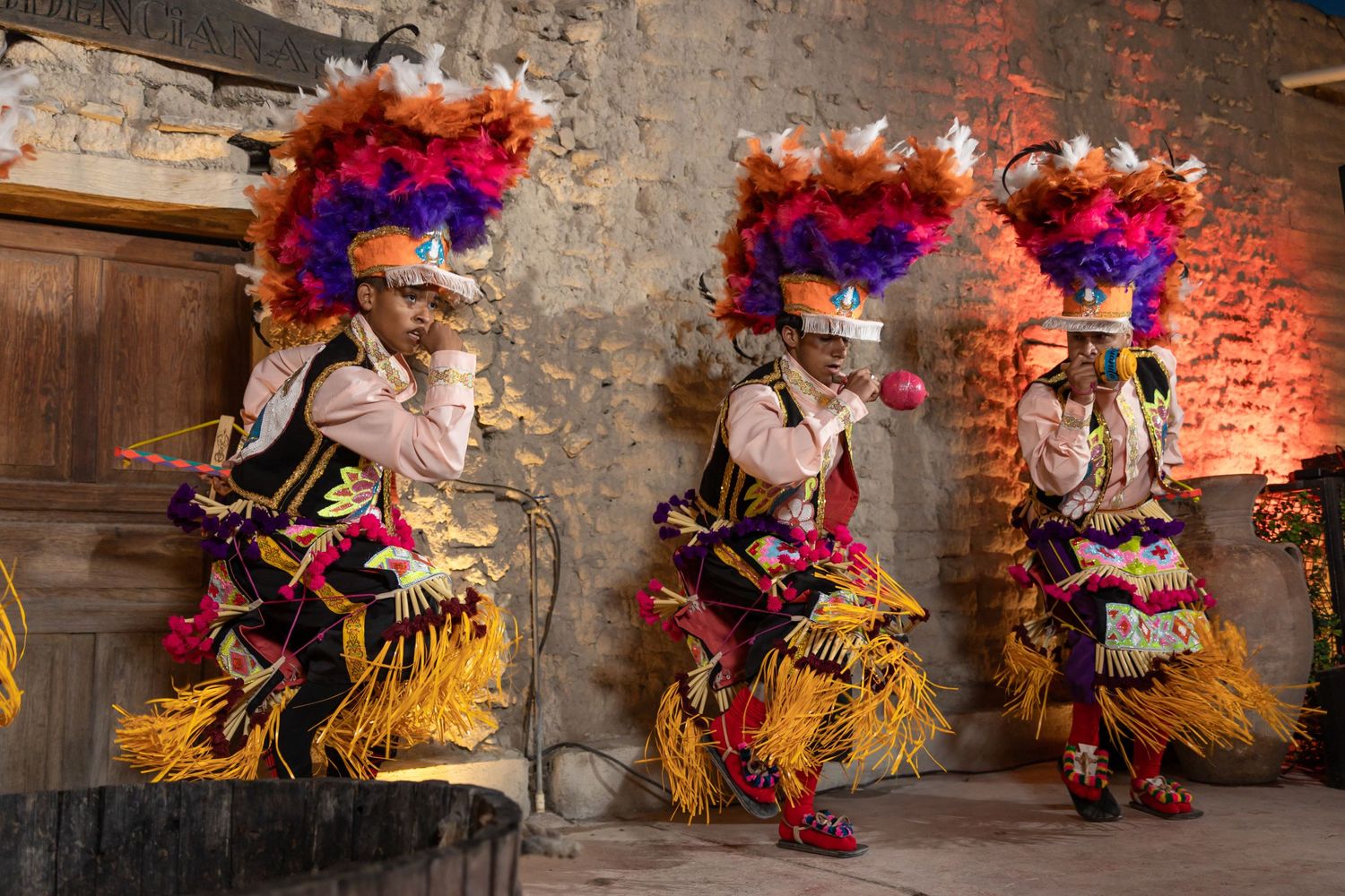 Performers in vibrant feathered costumes dance against a rustic stone wall background. Boda en Parras Coahuila fotógrafo