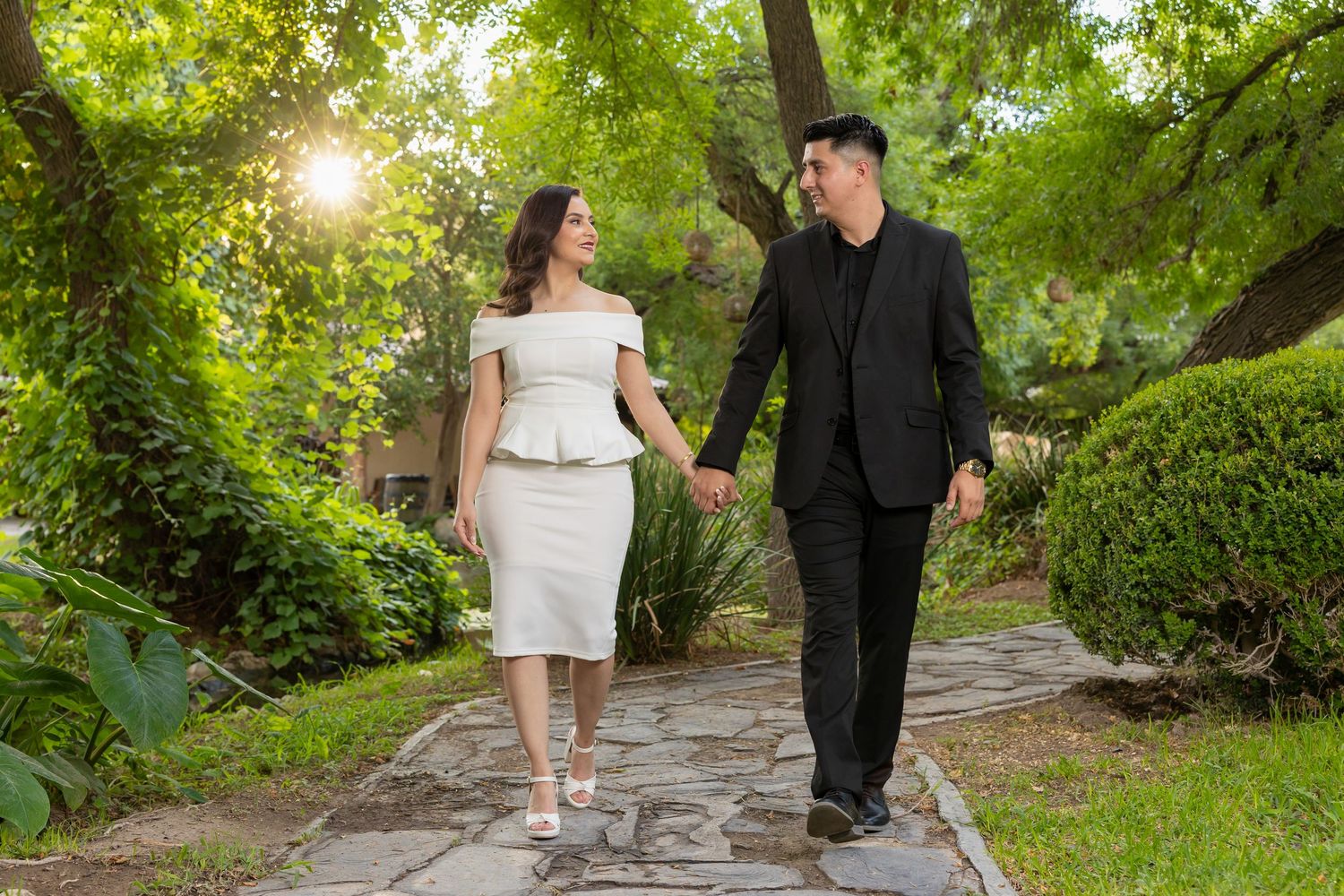 A couple walks hand in hand along a stone path in a lush garden with sunlight streaming through the trees.