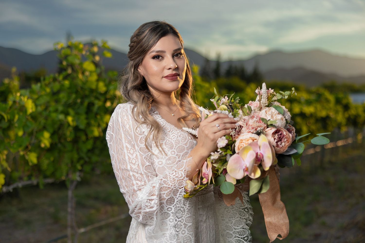 Woman in white lace dress holds delicate bouquet in vineyard at sunset against mountain backdrop. boda en Parras Coahuila fotógrafo