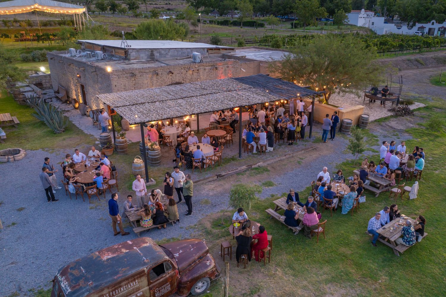 Aerial view of an outdoor gathering with people seated at tables under string lights at dusk.