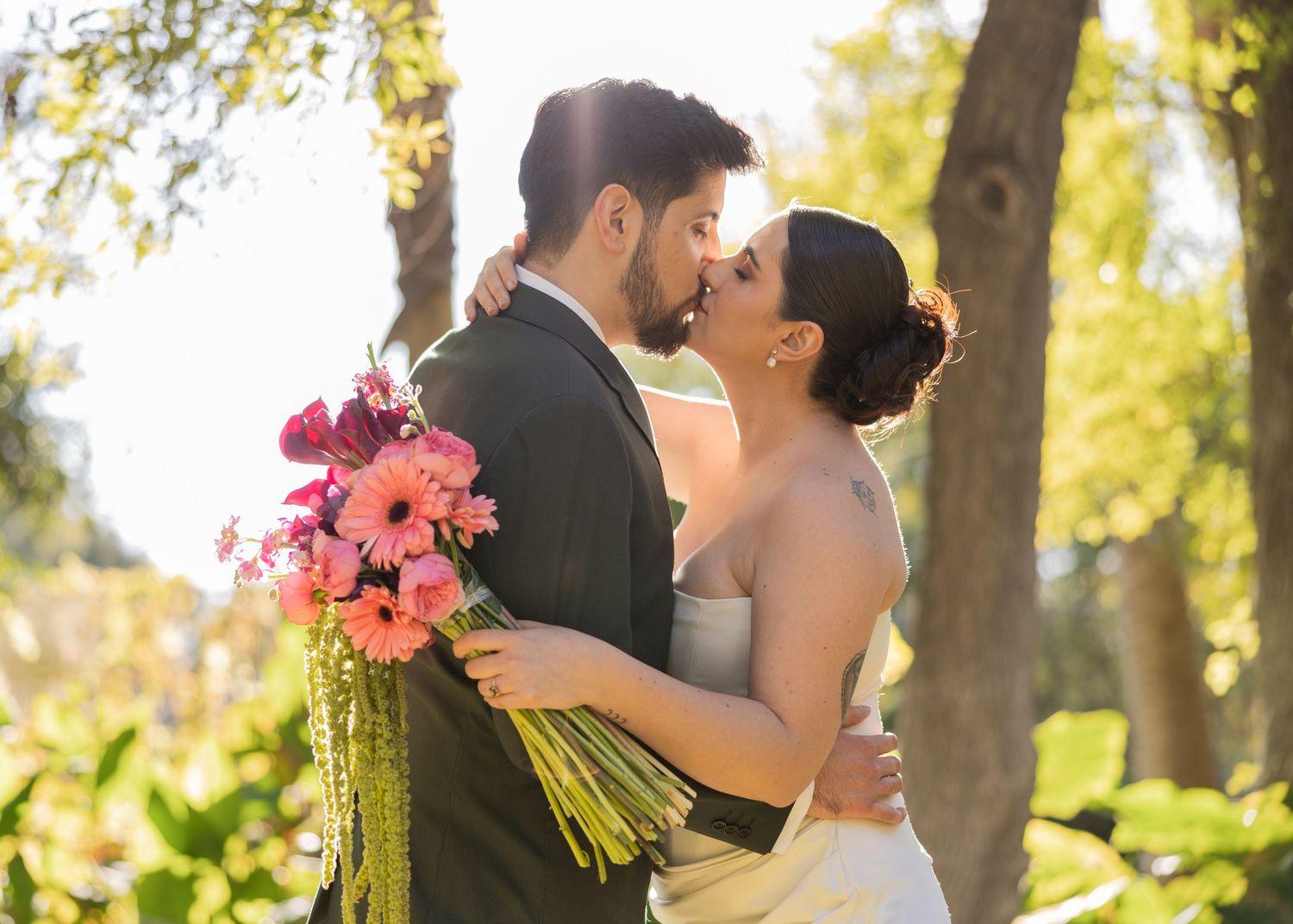A romantic wedding couple shares a tender kiss in a sunlit garden while holding a vibrant pink floral bouquet.