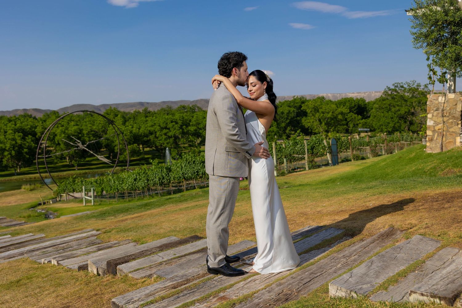 A couple shares a romantic moment in a scenic vineyard setting with rolling hills and blue skies in the background.