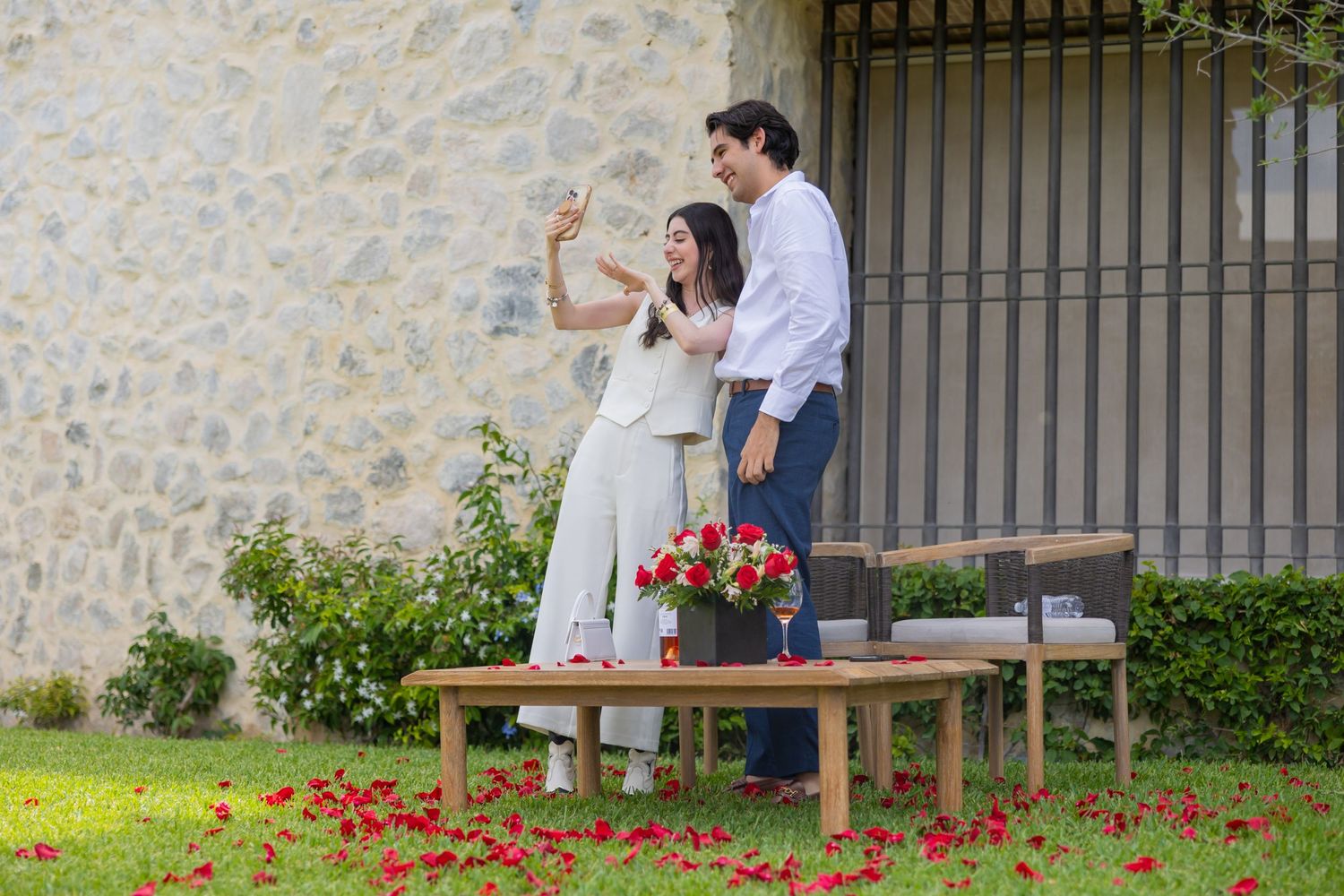 A couple celebrates on wooden benches with red rose petals scattered on the grass below. Propuesta de matrimonio Parras, Fotografo