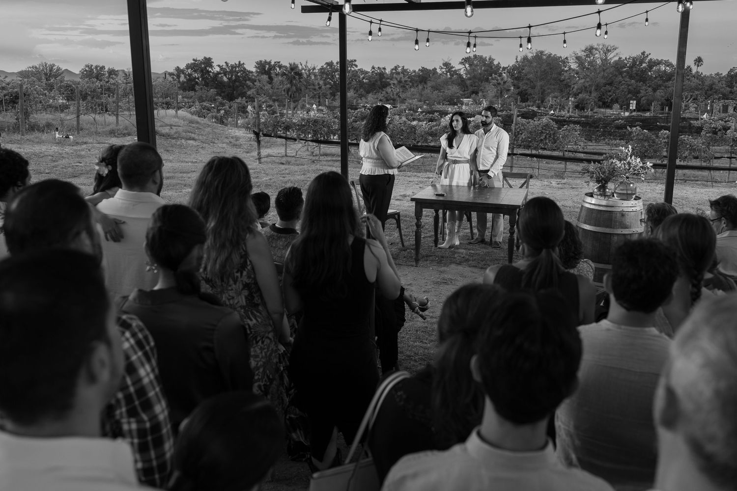 Black and white photo of a ceremony being conducted outdoors under string lights with guests watching. Boda en Parras Coahuila fotógrafo