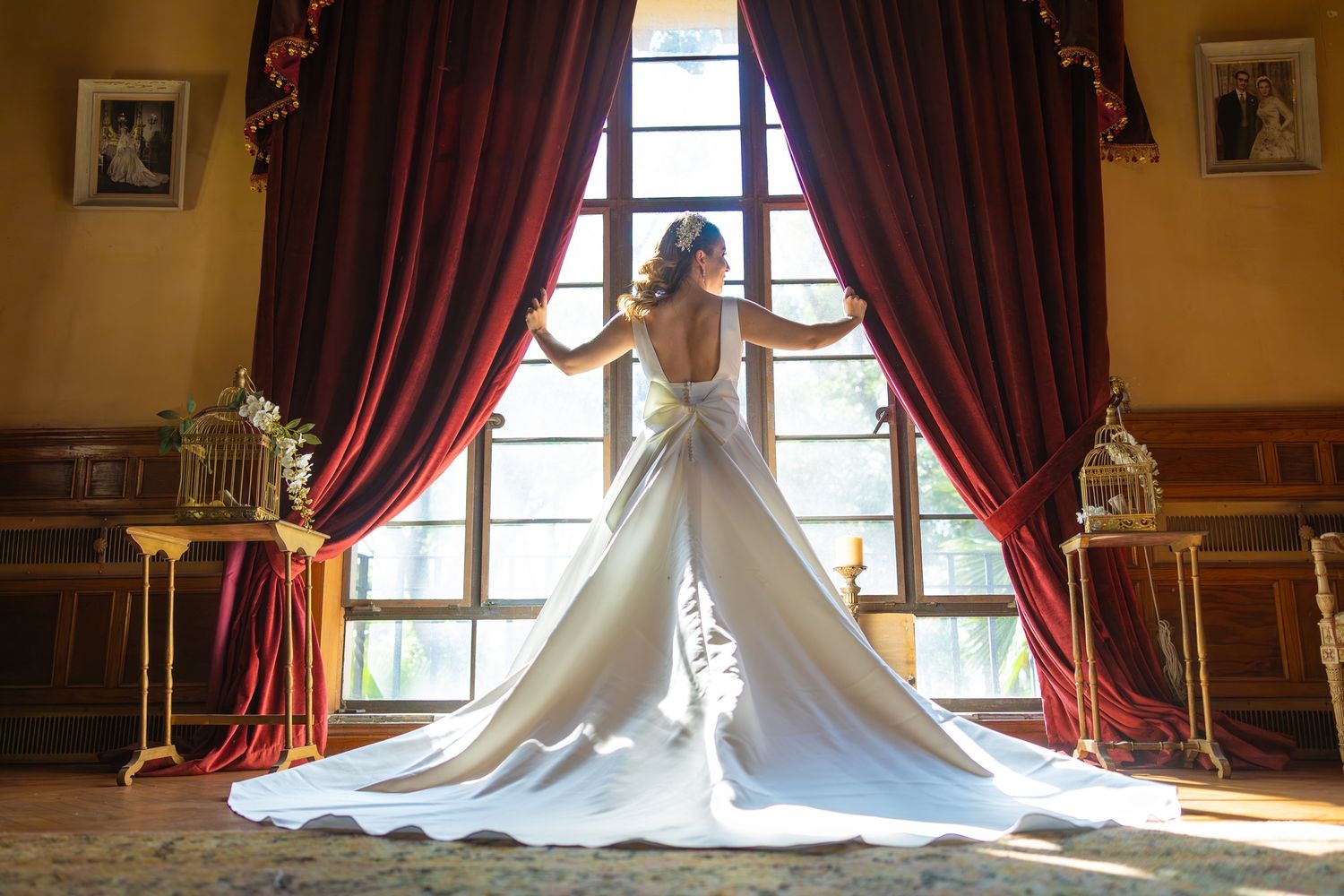 Elegant wedding dress with long train photographed against dramatic red curtains and window in ornate room. boda en Parras Coahuila fotógraf