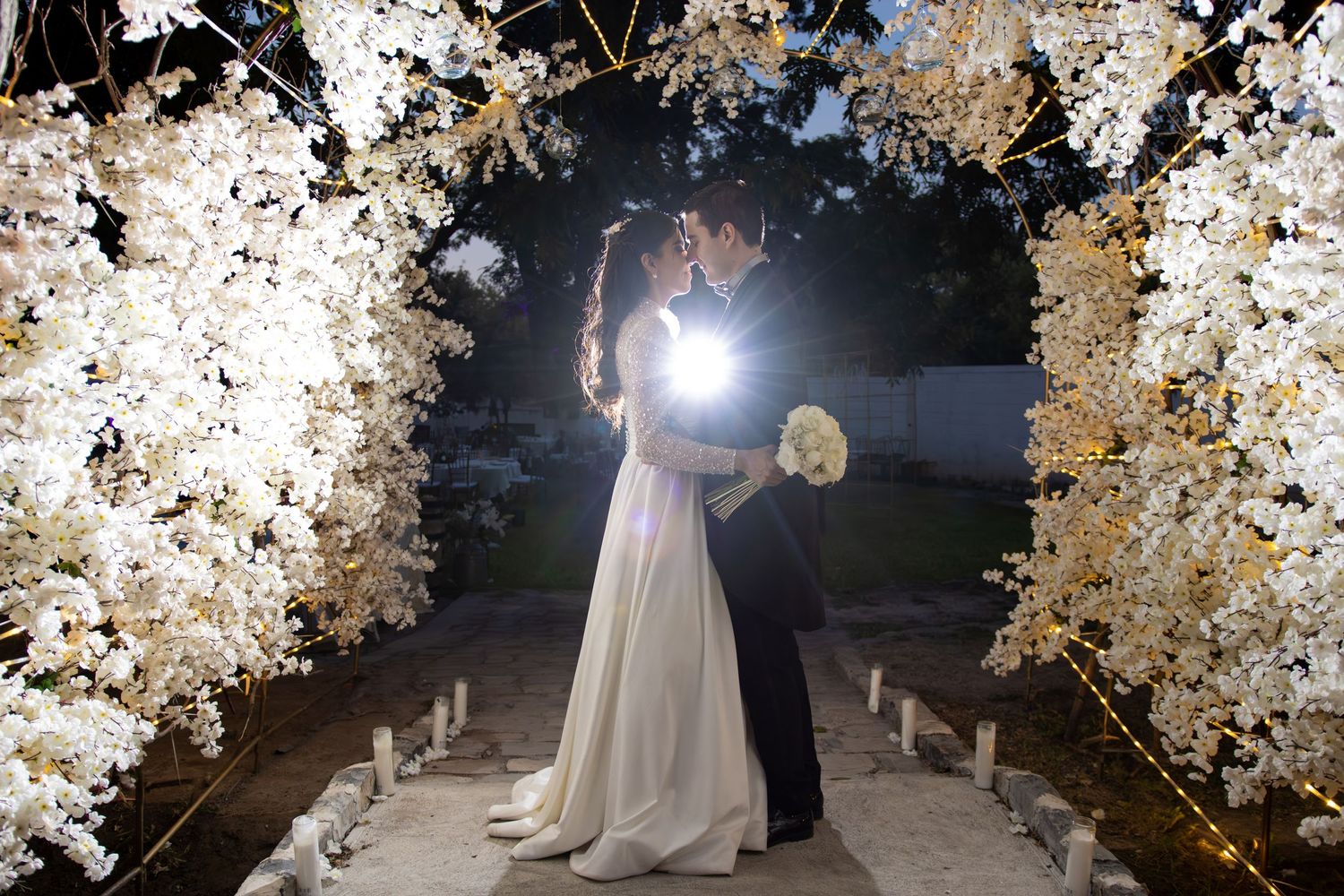 A wedding couple embraces under a stunning floral arch with white blossoms and dramatic lighting at night. Boda en Parras Coahuila, Foto