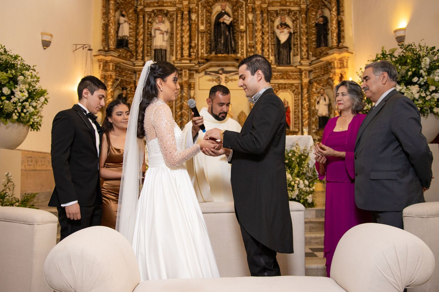 A wedding ceremony takes place in front of an ornate golden altar with attendants watching. Boda en Parras Coahuila, Fotógrafo de bodas