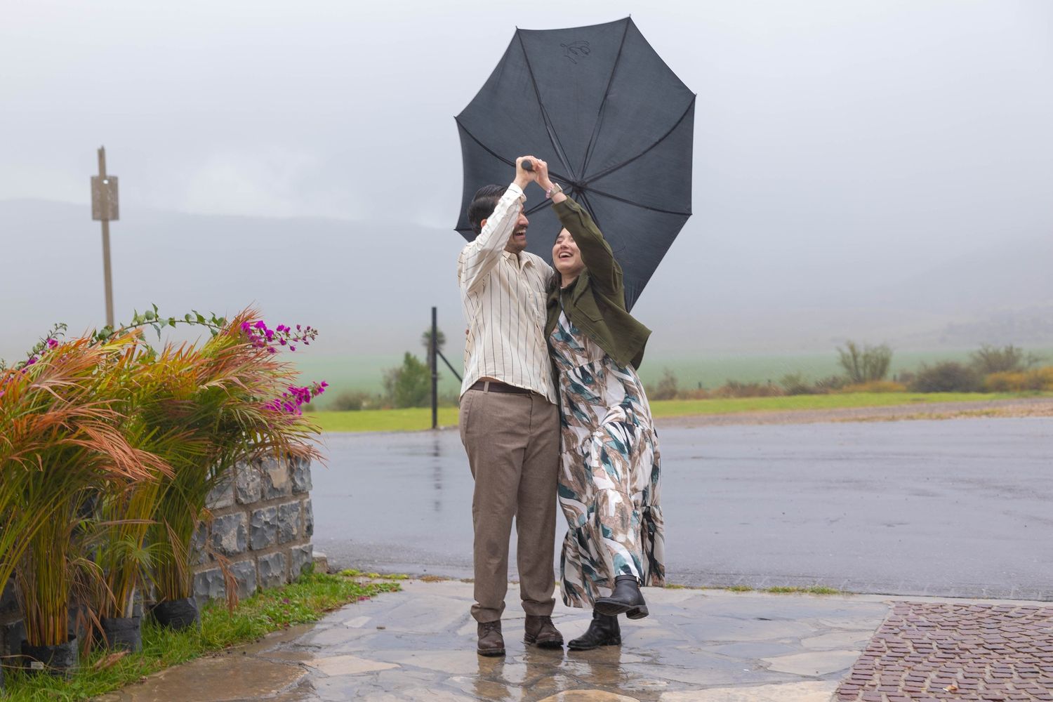 Couple dancing playfully with black umbrella on rainy day surrounded by tropical plants. boda en Parras Coahuila fotógrafo
