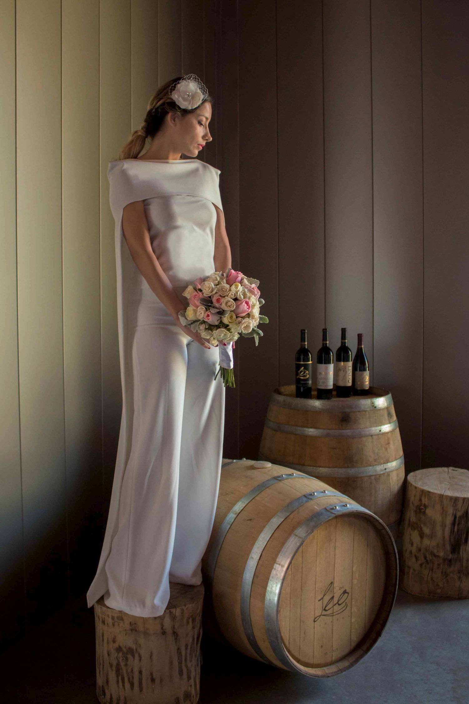 Elegant bride in simple white dress holding bouquet next to wine barrels in rustic setting. boda en Parras Coahuila fotógrafo