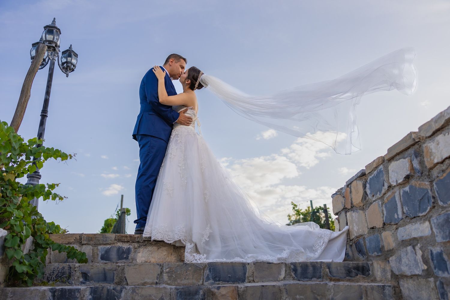 Couple in wedding attire poses on stone steps at sunset with ornate iron lamp post and architecture in background.
