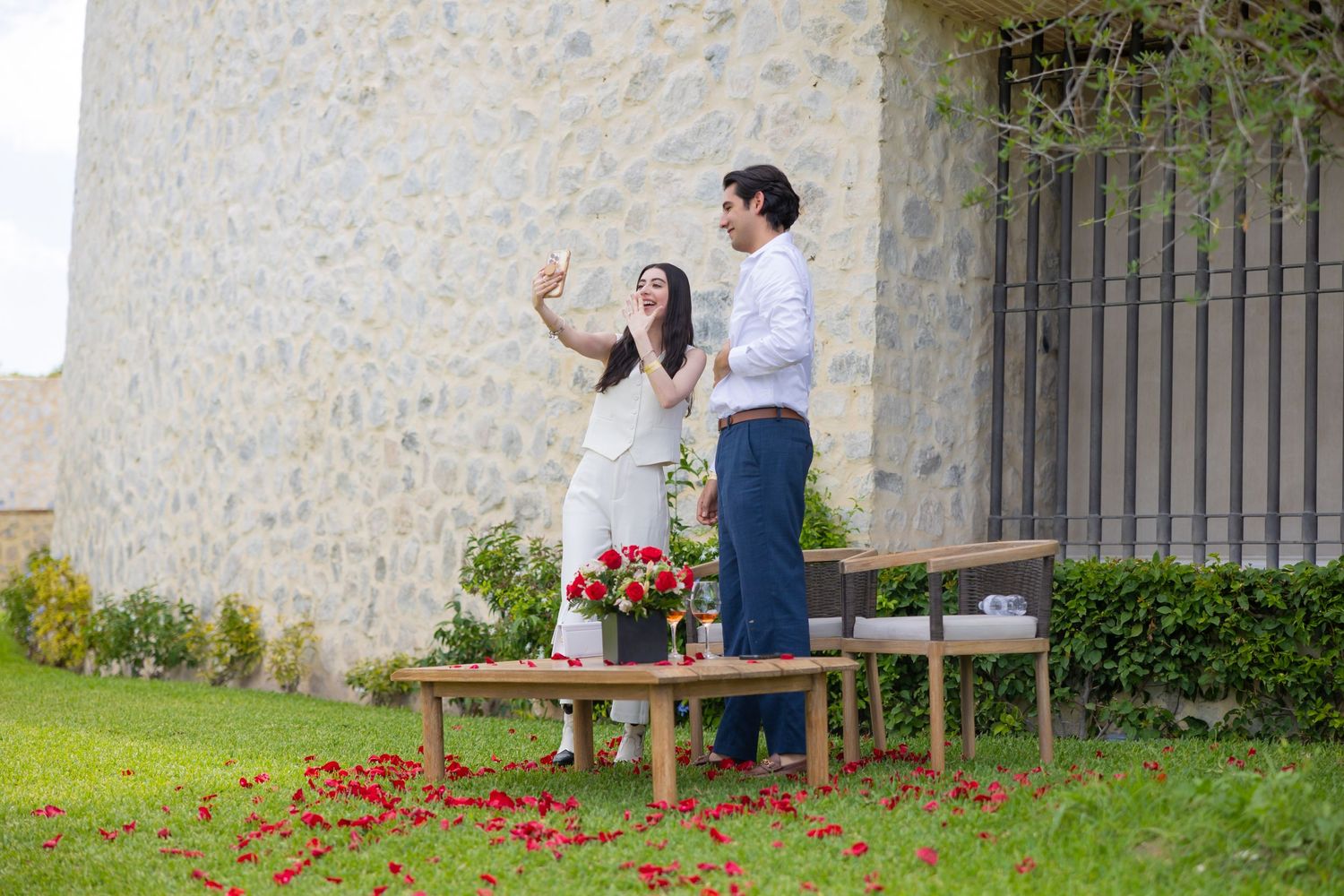 Two people stand on wooden benches against a stone wall backdrop with red petals scattered below. Propuesta de matrimonio Parras, Fotografo