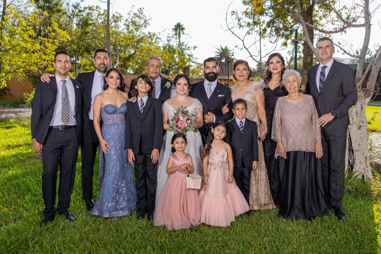 A formal family wedding portrait with multiple generations posed outdoors under trees. Boda en Parras Coahuila, Fotógrafo de bodas