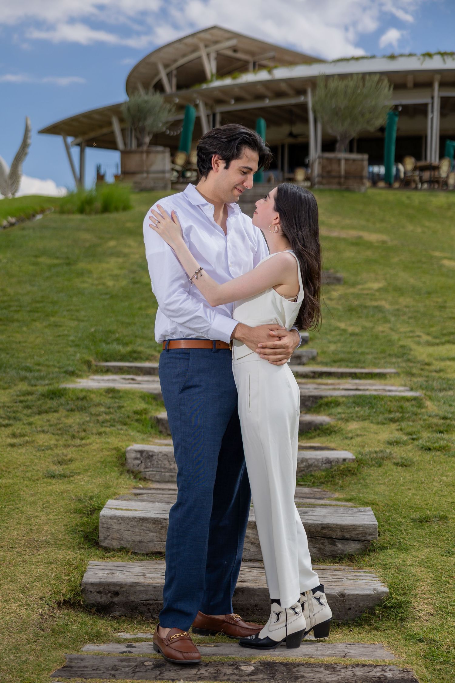 A couple embraces on stone steps with a modern building and green hillside behind them. Propuesta de matrimonio Parras, Fotografo