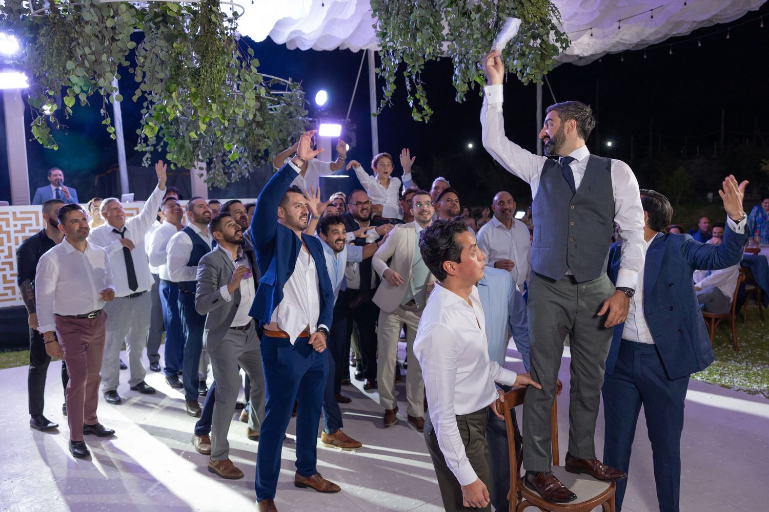 Celebration scene with guests dancing and cheering under illuminated tent with hanging greenery. Boda en Parras Coahuila fotógrafo