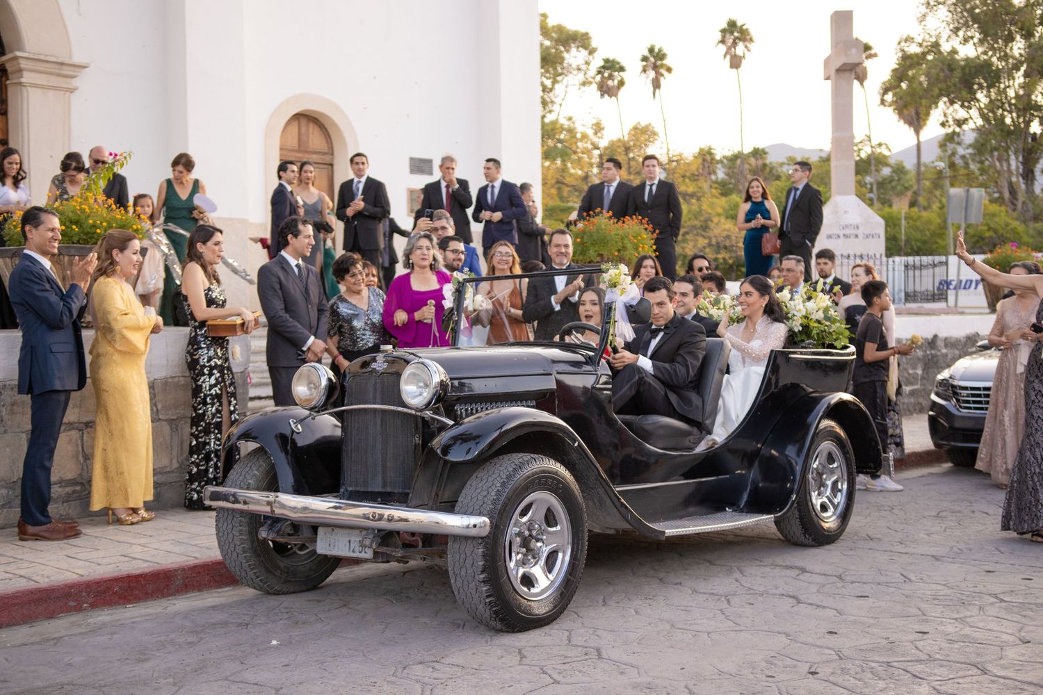 Classic black vintage car parked outside church with wedding guests gathering around. Boda en Parras Coahuila, Fotógrafo de bodas