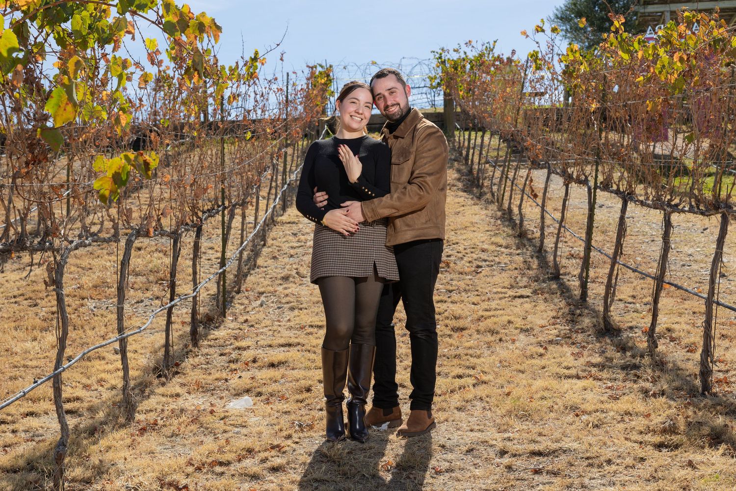 A couple poses together in a scenic vineyard during autumn with bare grapevines and golden foliage in the background.
