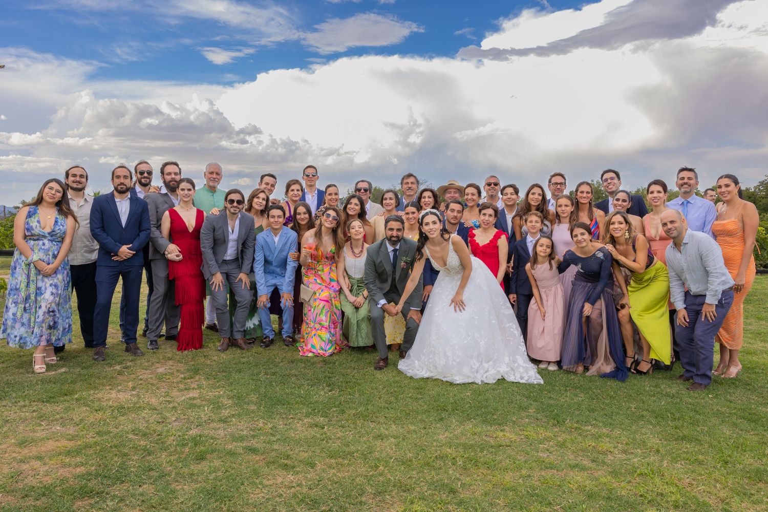 Large wedding party group poses together on a grassy hill under blue skies with white clouds. Boda en Parras Coahuila fotógrafo