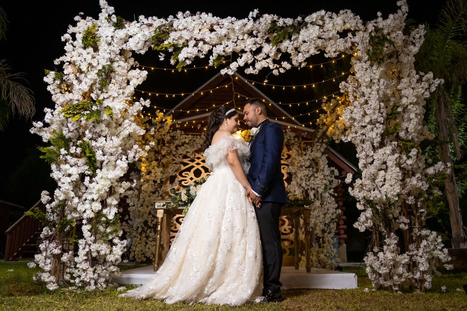 Newlyweds pose under ornate white floral arch with twinkling lights at evening wedding. boda en Parras Coahuila fotógrafo