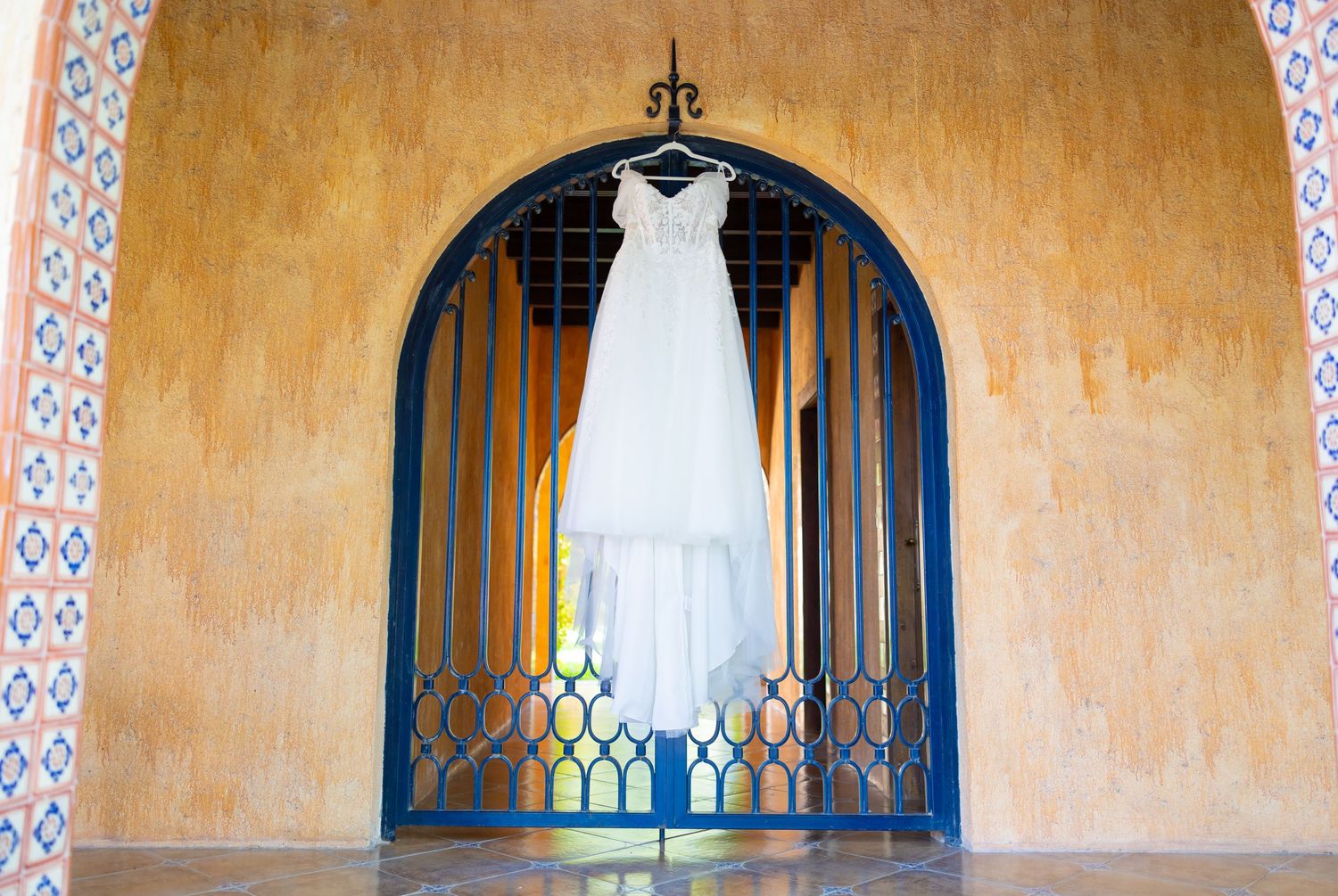White wedding dress hanging in arched Spanish-style window with ochre wall. Boda en Parras Coahuila, Fotógrafo de bodas