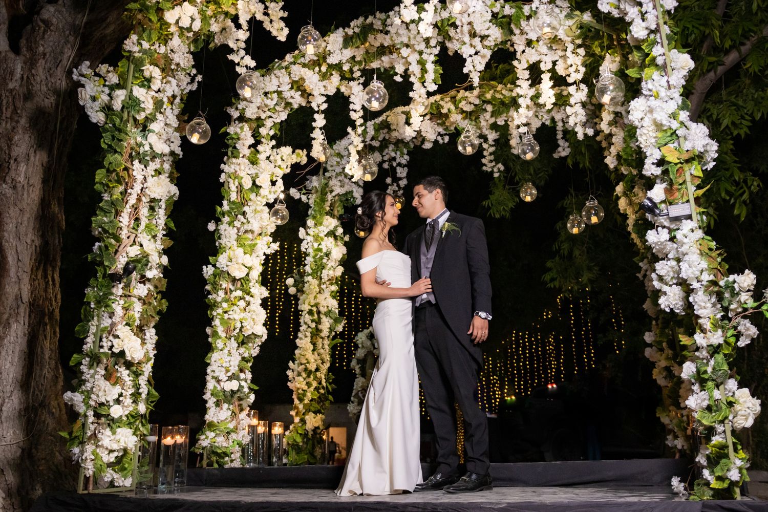 A romantic wedding ceremony under a beautiful floral arch with white orchids and greenery in an outdoor garden setting.