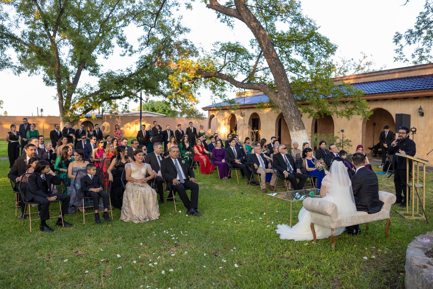 Outdoor wedding ceremony with guests seated on a lawn near Spanish-style buildings at sunset. Boda en Parras Coahuila, Fotógrafo de bodas