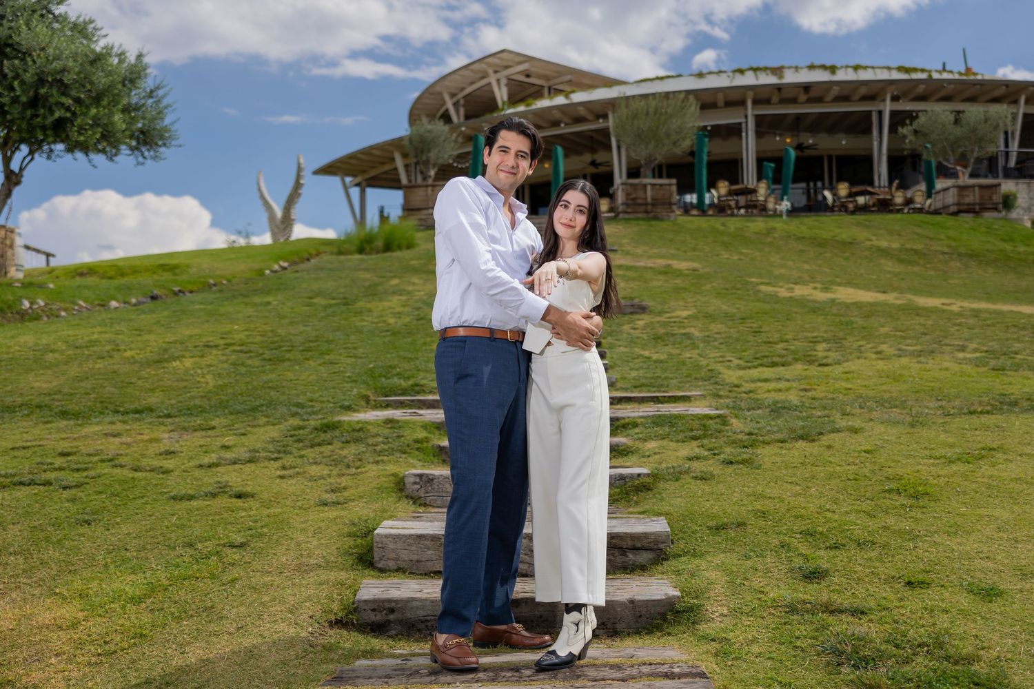 A couple stands together on stone steps with a modern venue and blue sky in the background. Propuesta de matrimonio Parras, Fotografo