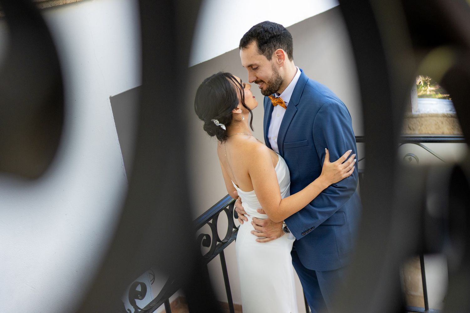 A romantic moment between newlyweds in formal attire captured through an ornate railing at their wedding celebration.