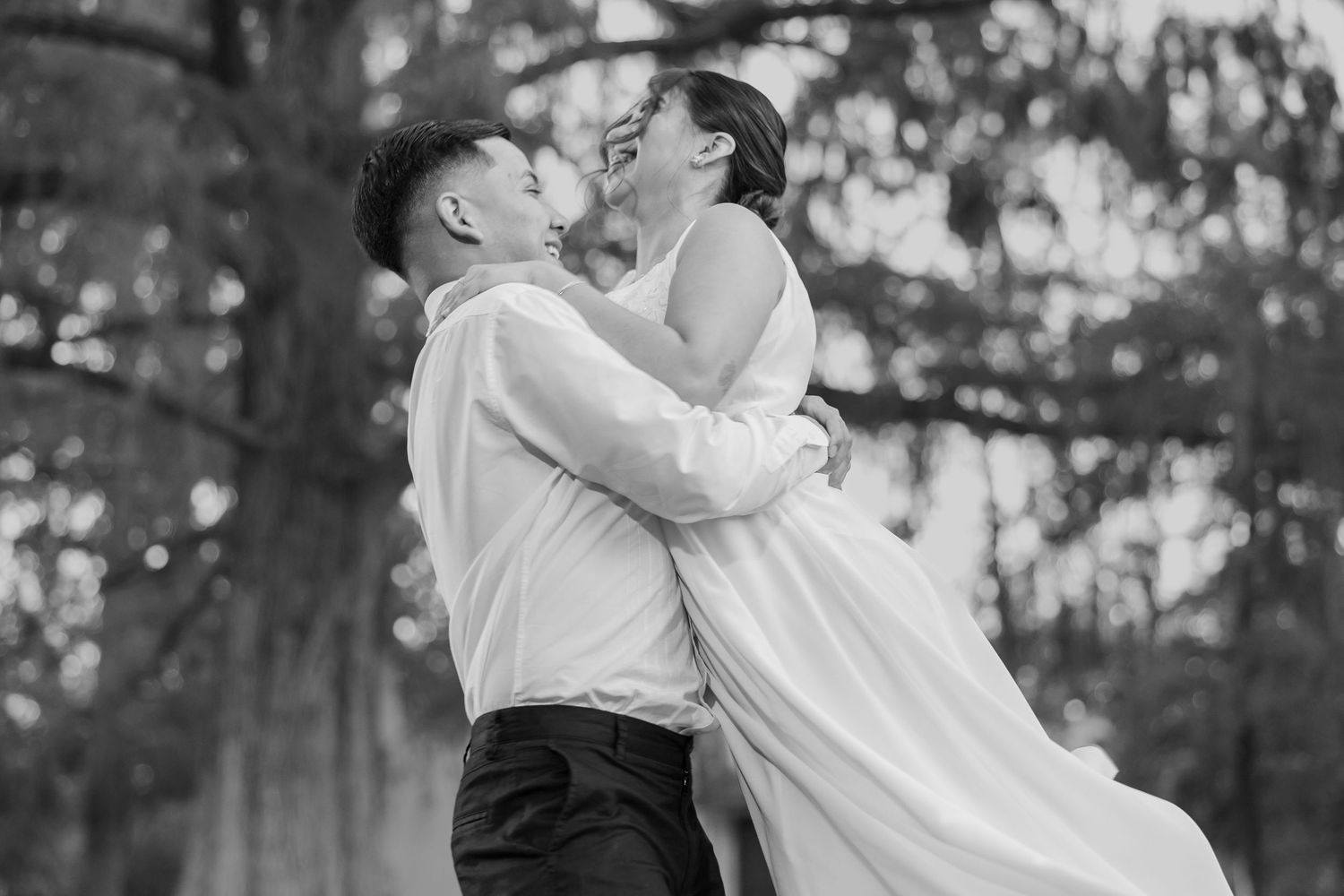 A romantic couple in wedding attire embraces and kisses in a dreamy outdoor setting with natural light filtering through trees.