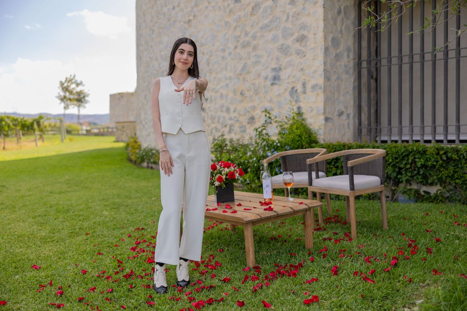 Woman stands beside outdoor picnic setup with wine and flowers against stone wall background. Propuesta de matrimonio Parras, Fotografo