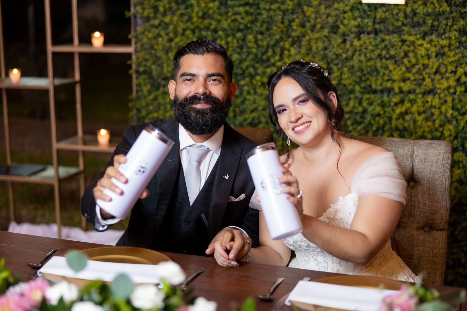 Wedding couple sits at decorated table holding matching drink tumblers during outdoor reception. Boda en Parras Coahuila, Fotógrafo de bodas