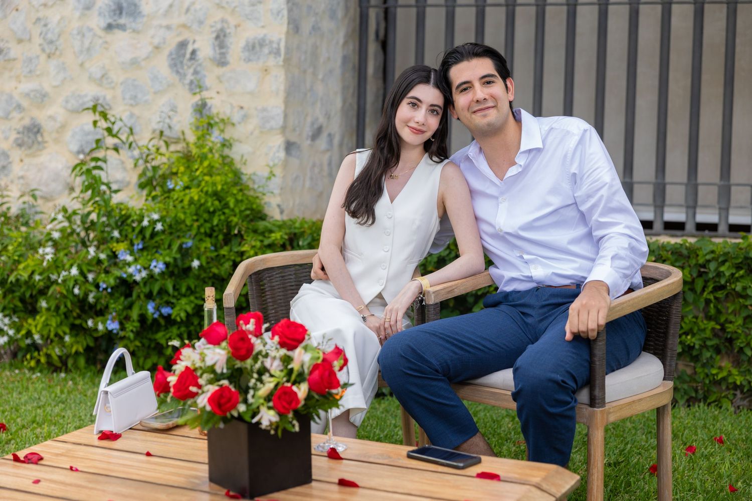 Couple seated on bench with red flower arrangement in garden setting. Propuesta de matrimonio Parras, Fotografo