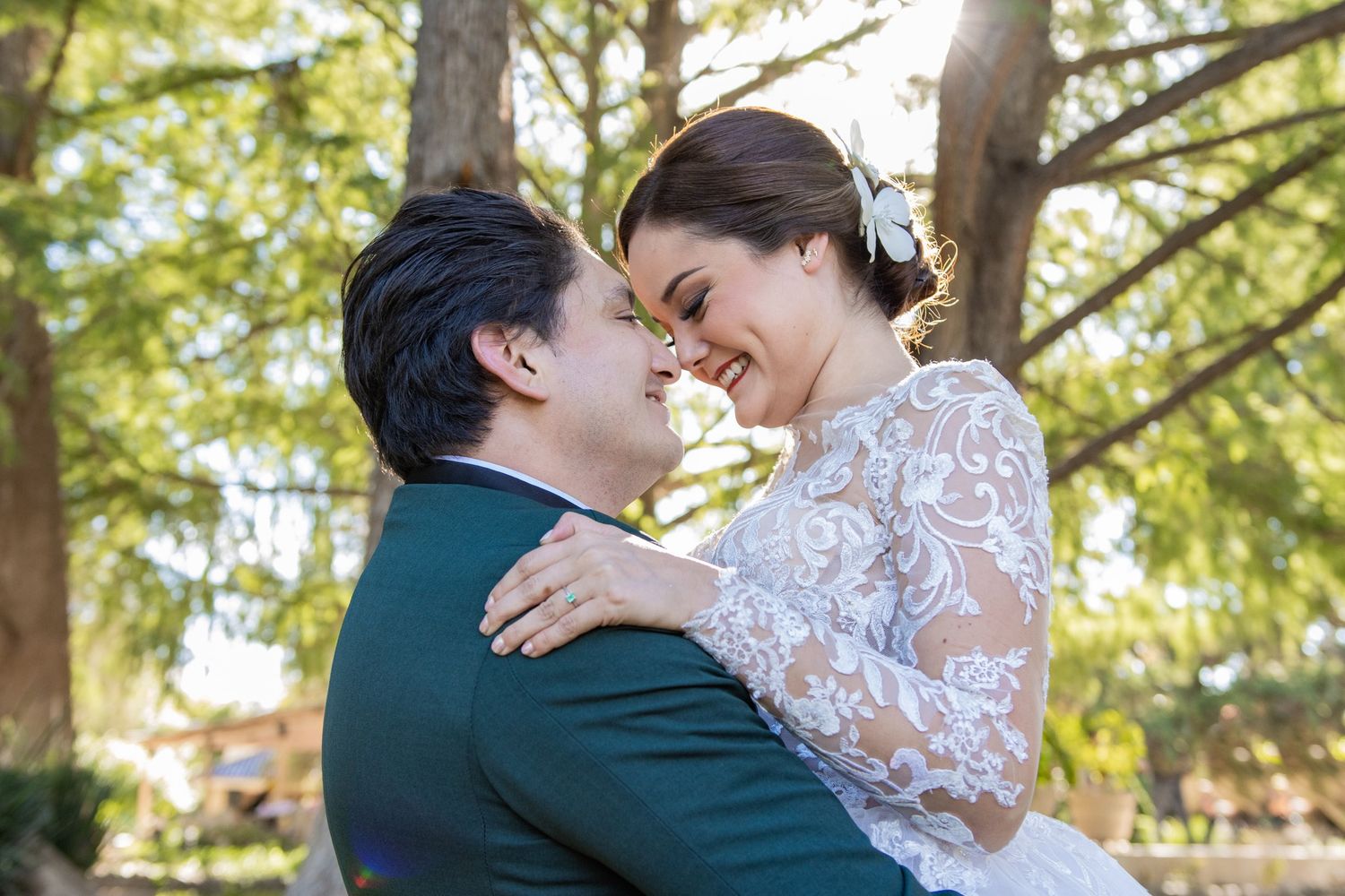 A romantic couple in wedding attire embrace outdoors with sunlit trees in the background.