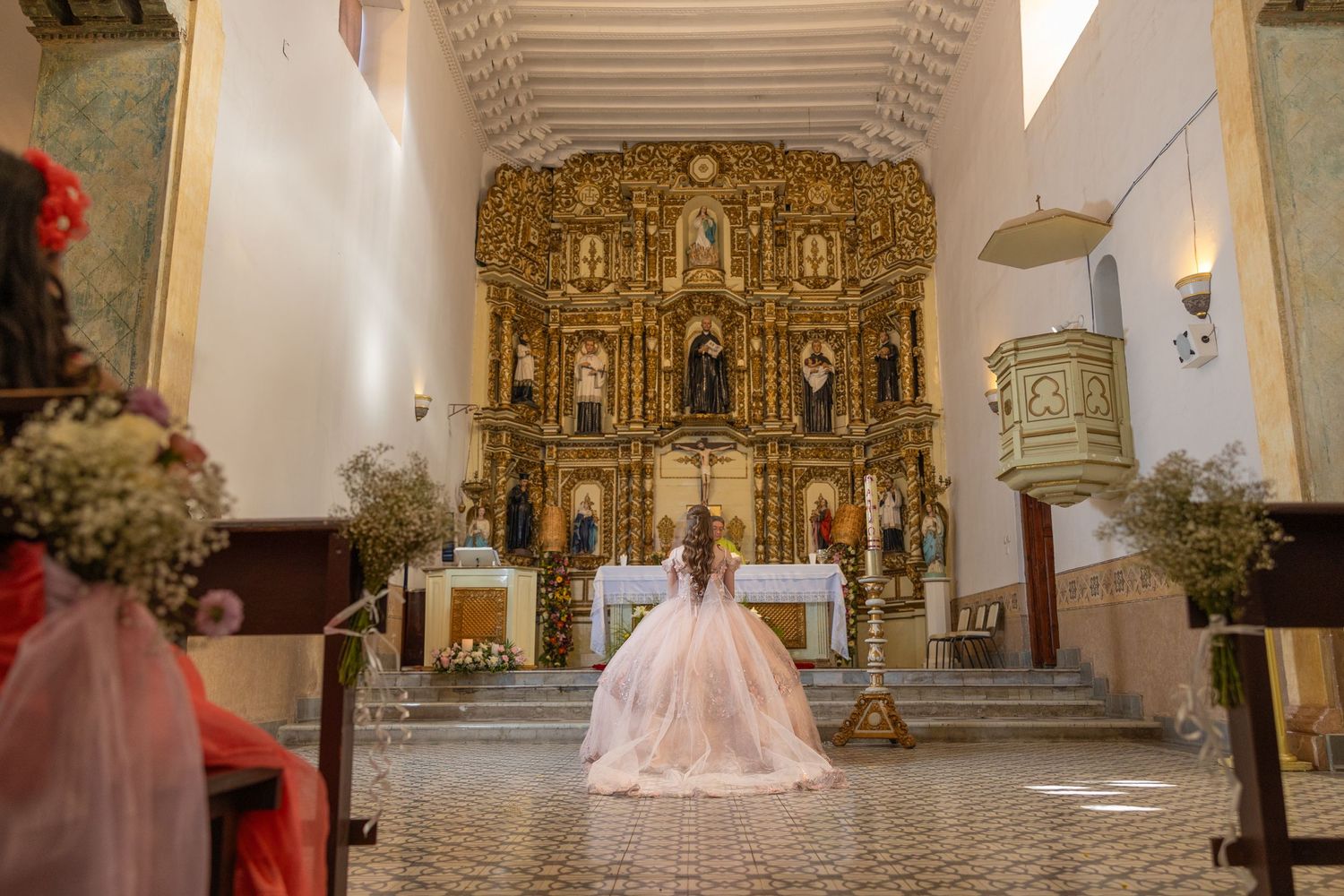 A dramatic full-length shot of a pink ball gown in a church with an ornate golden altar backdrop. Evento en Parras Coahuila , Fotógrafo Rulo