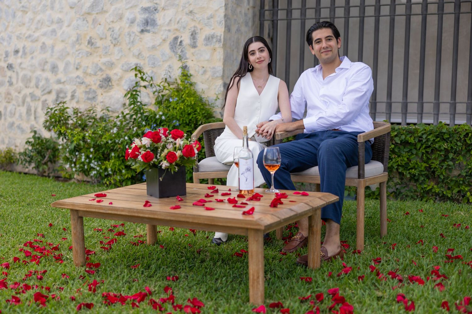 A couple relaxes on outdoor furniture with wine and snacks among red flowers. Propuesta de matrimonio Parras, Fotografo