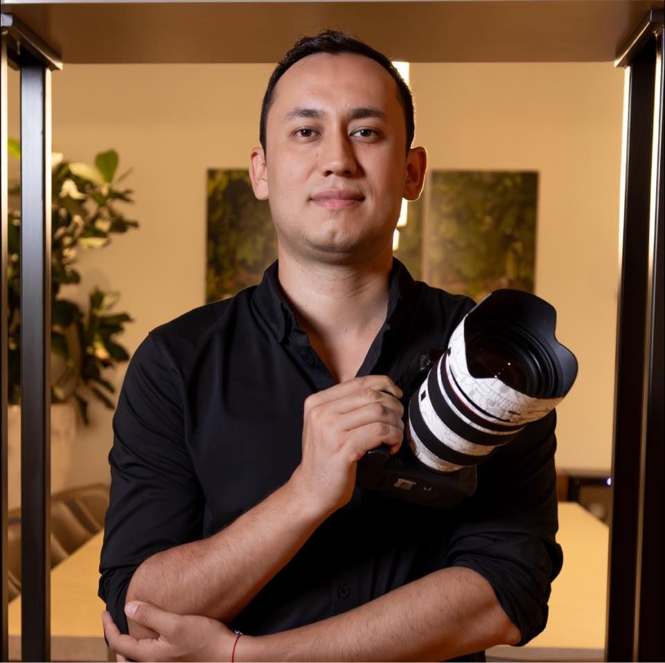 Professional photographer in black shirt holding a camera with telephoto lens in studio setting with plants in background.