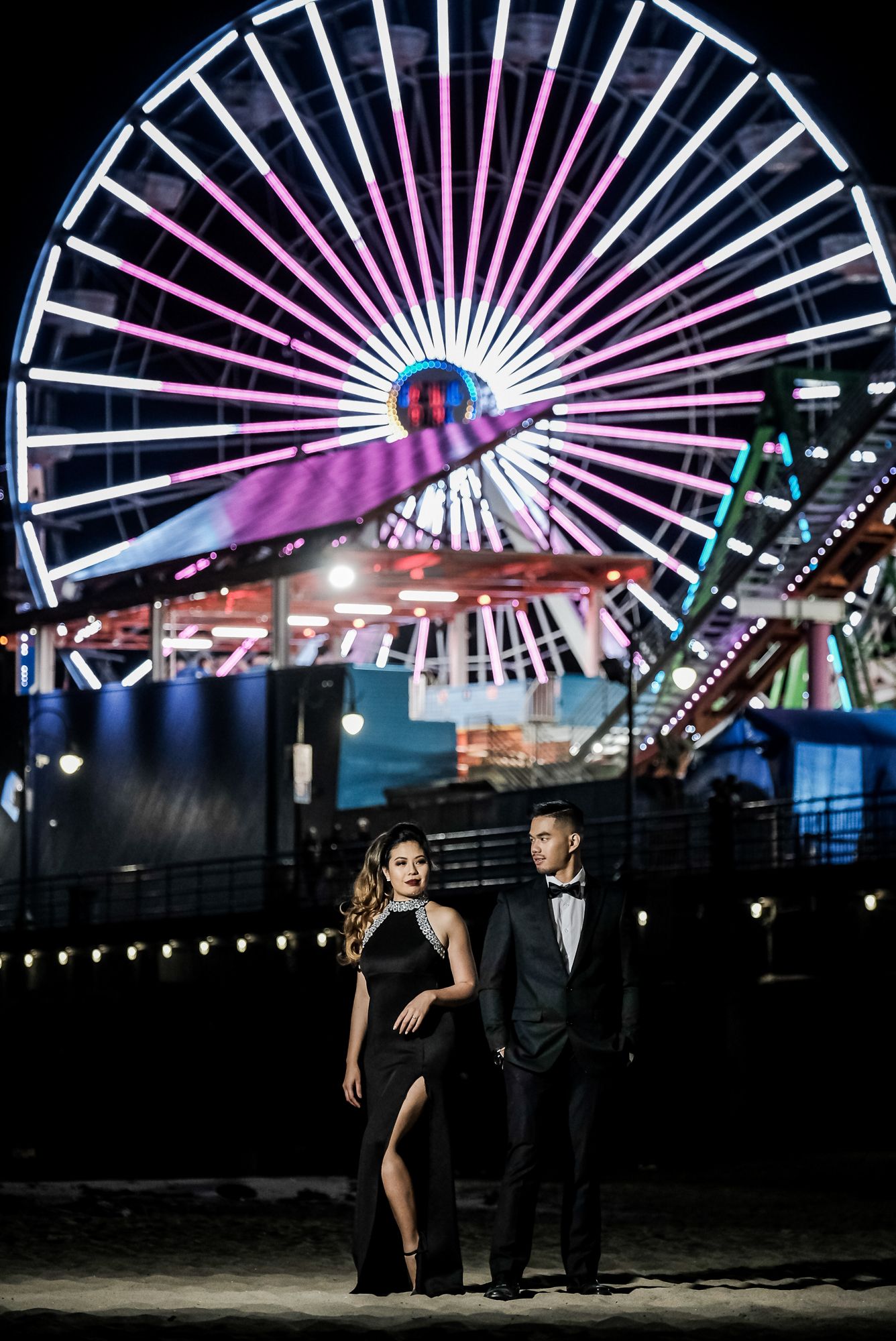 Couple in formal attire posing in front of illuminated Ferris wheel at Santa Monica Pier at night.