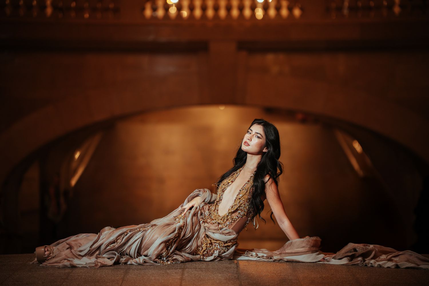 Elegant woman in a flowing gold gown posing gracefully on a wooden floor in a warmly lit venue.