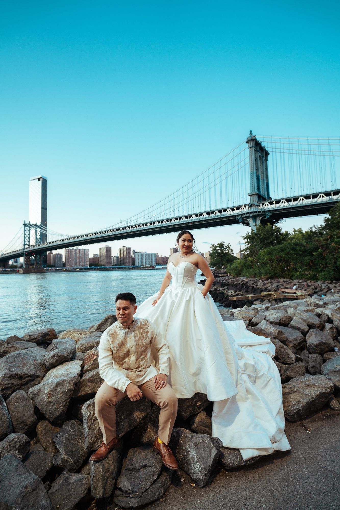 Bride and groom posing on rocky shore with Manhattan Bridge and NYC skyline in background.