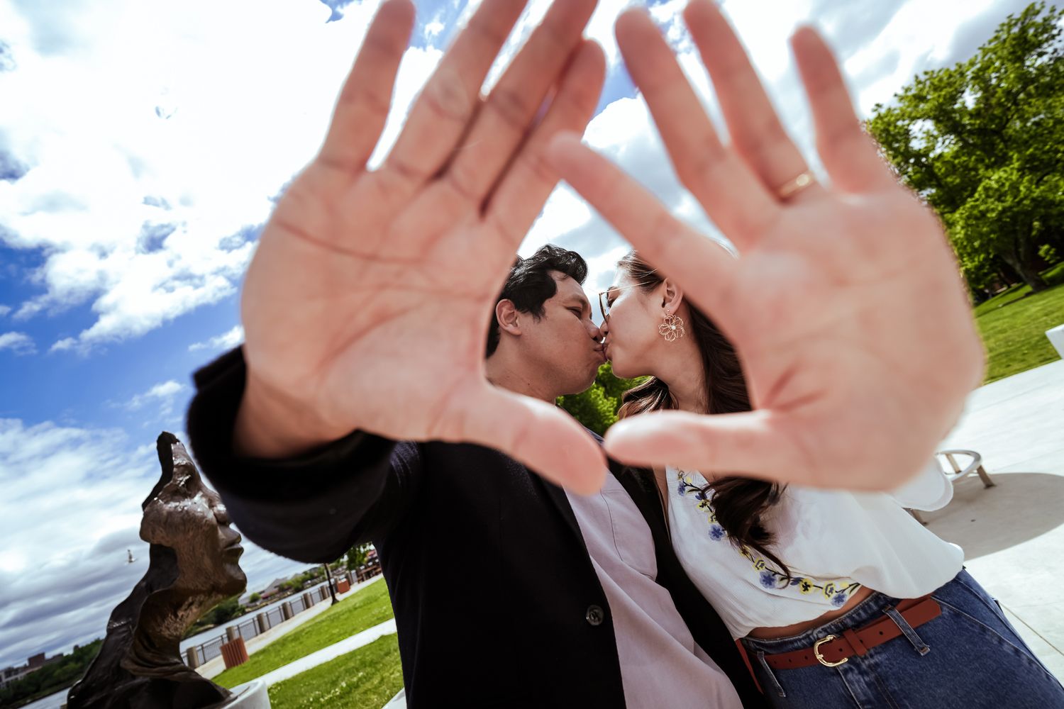 Couple kissing behind outstretched hands framing the shot outdoors near a bronze statue.