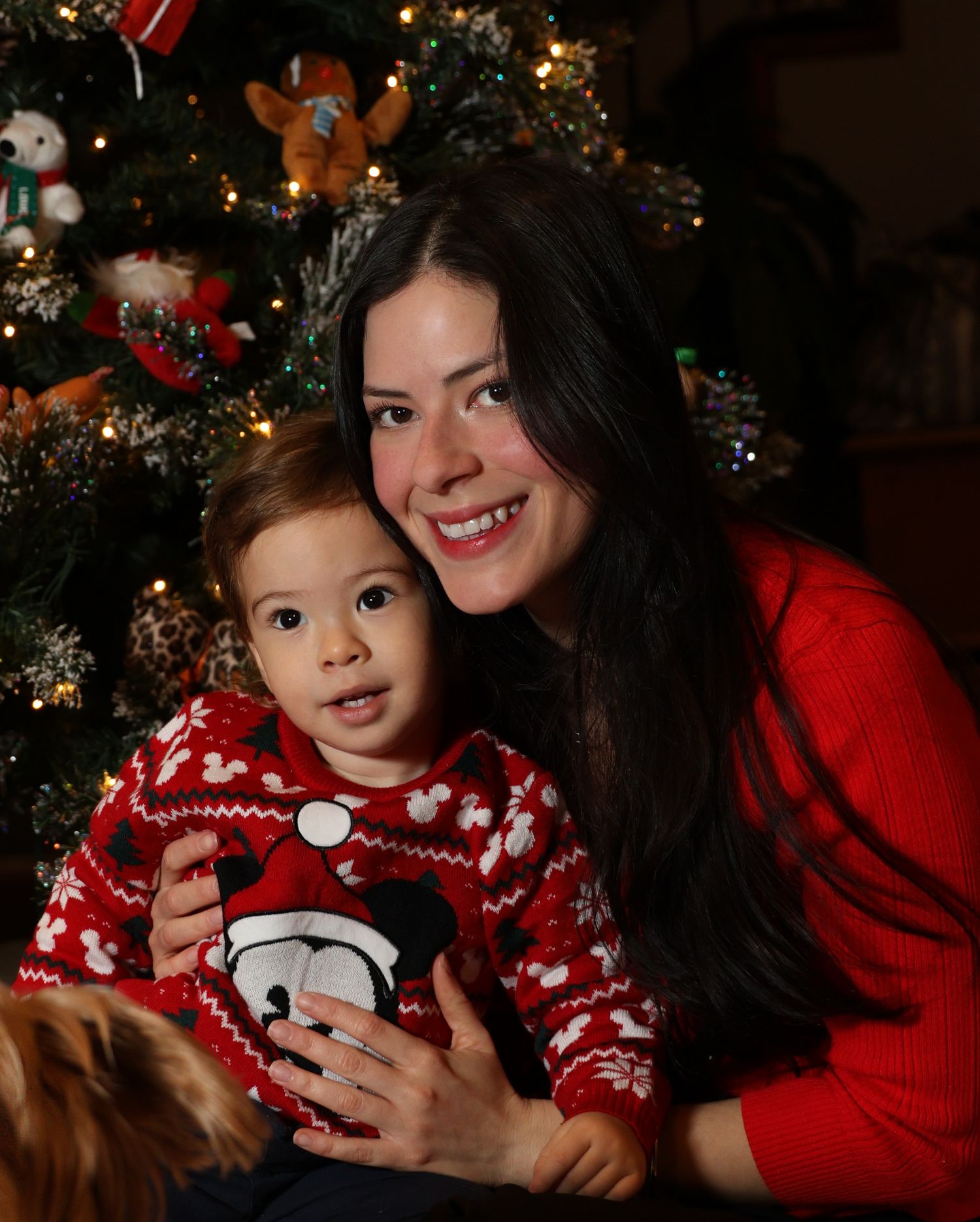 Mother and child in matching red holiday sweaters pose by Christmas tree in warm lighting.