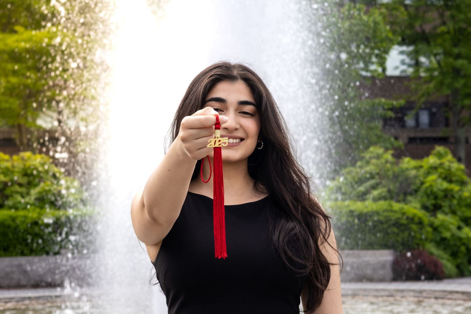 Someone in a black shirt holds up a graduation tassel while posing in front of a water fountain on a sunny day.