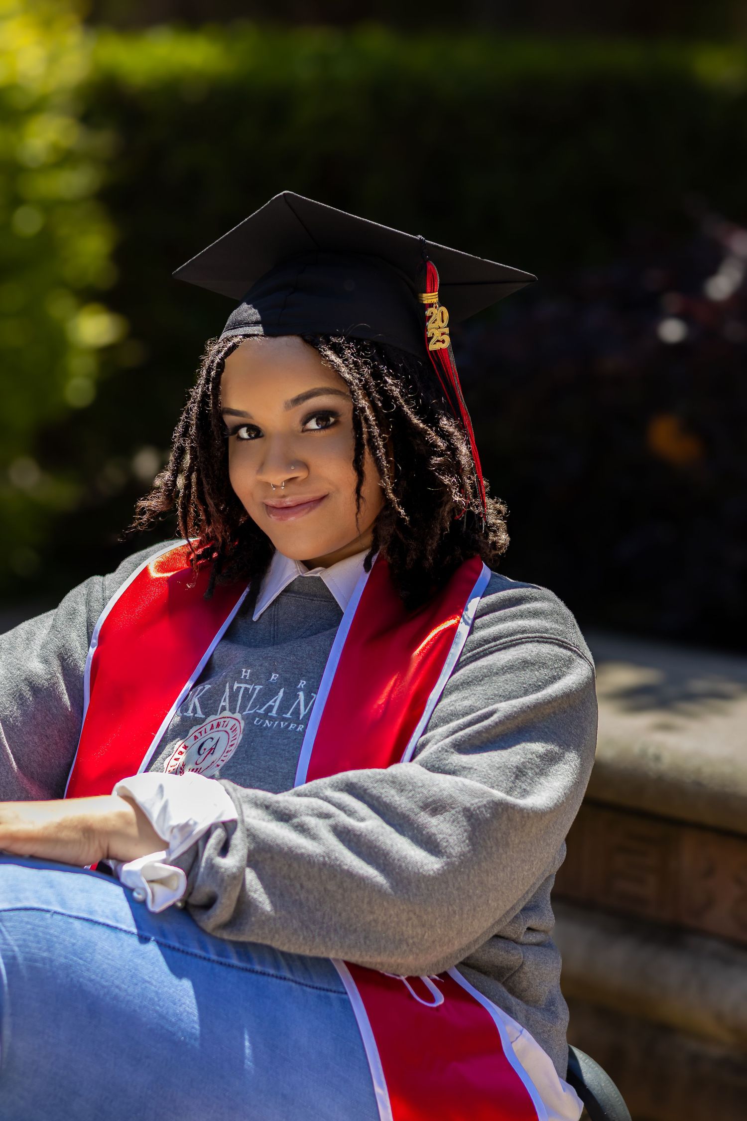 Graduate in cap and gown wearing red stole and grey sweatshirt sits outdoors for graduation portrait with natural lighting.