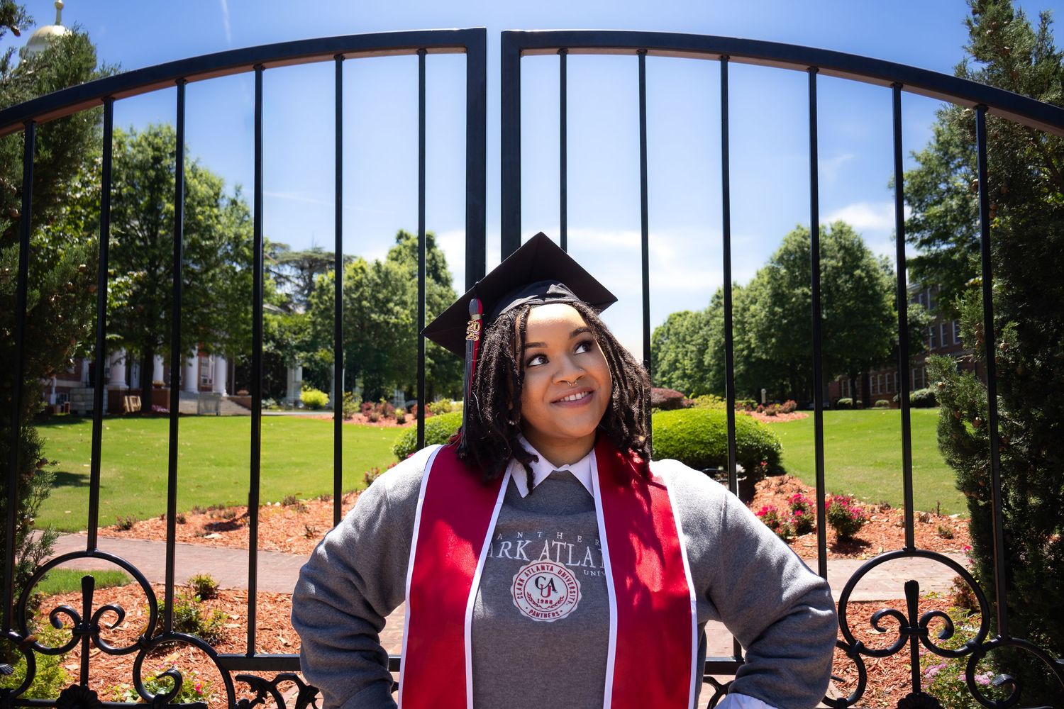Graduate in cap and gown stands in front of decorative iron gate at university with green campus lawn visible behind.