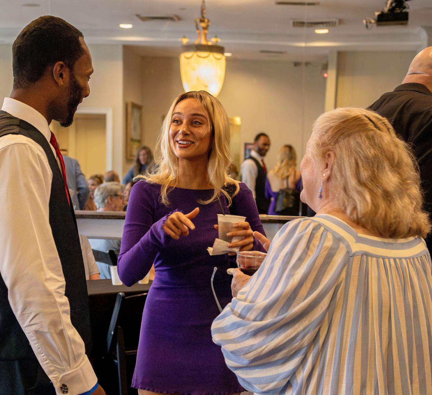People in formal attire dance and socialize at an indoor event with warm lighting and purple accents.