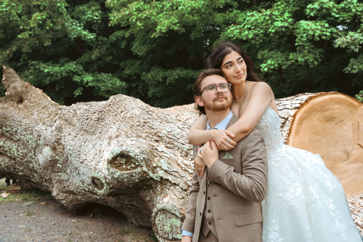 Bride and groom posing romantically near a fallen tree trunk surrounded by lush green foliage in an outdoor wedding photo.