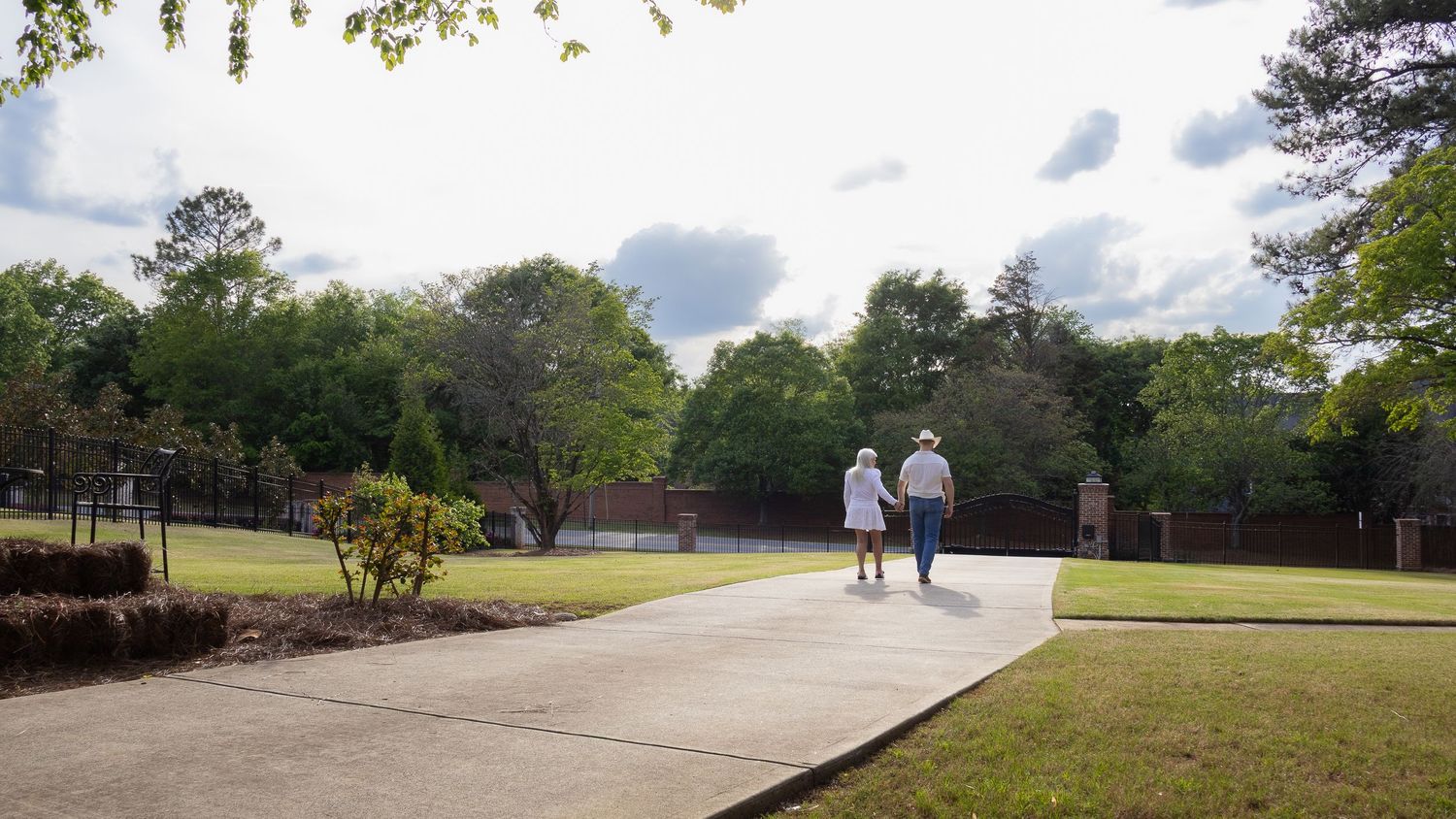 Two people walk together on a paved pathway through a lush park landscape on a partly cloudy day.